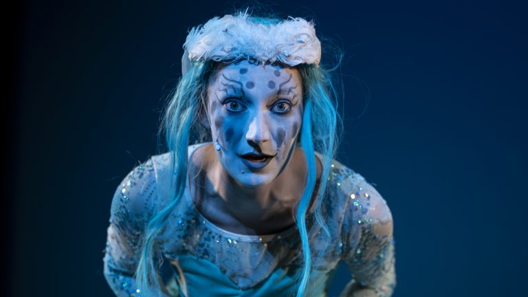 ALT-TEXT_ A ballerina dressed in_ a blue tutu, blue hair, and feathered crown, is staring into the camera lens. -Photo by Marc J